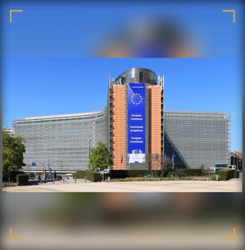 An exterior view of the Berlaymont building in Brussels, headquarters of the European Commission, featuring its distinctive curved glass facade and a central tower displaying a large blue banner with EU stars.