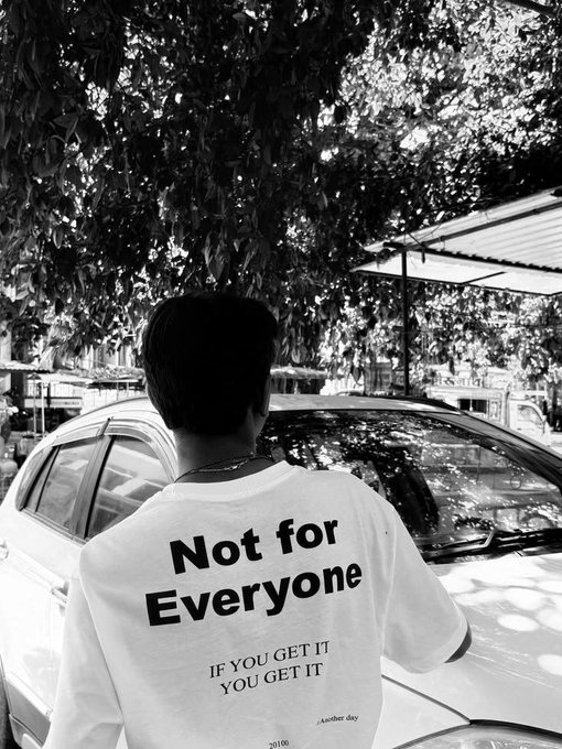 A black and white photo of a man seen from behind, wearing a white t-shirt with the text 'Not for Everyone' and 'IF YOU GET IT YOU GET IT' printed on the back. He stands near a car under a leafy tree.