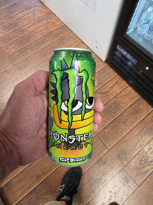 A hand holds up a can of Monster Energy 'Slim Ducks Taurine' featuring a green crocodile/monster design, photographed above wooden flooring with a vending machine in the background.