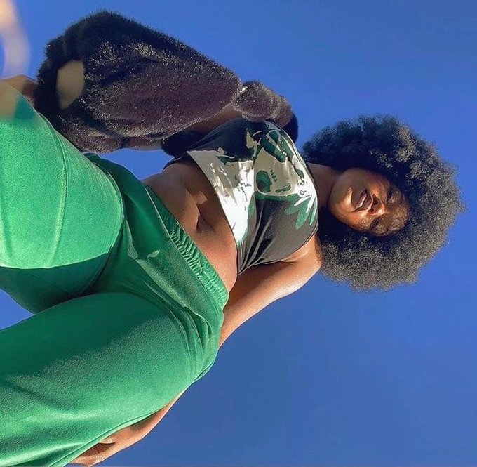 The image features a woman with long hair wearing green pants, standing against a blue sky. She has her arms outstretched towards the camera, giving a close-up view of herself. There are no texts or other objects present in the image.