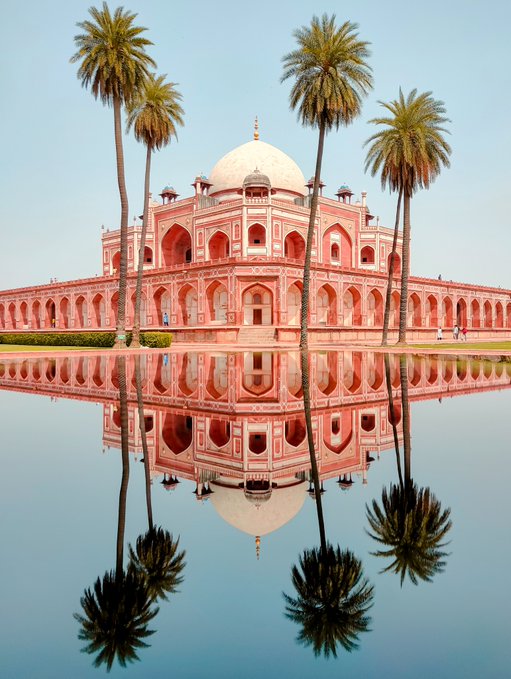 A red sandstone building with a central white dome and arched colonnades, flanked by tall palm trees, is perfectly reflected in a still pool of water beneath it.