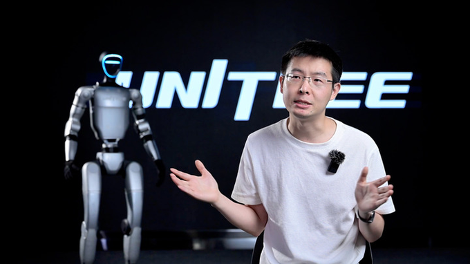 A man in a white t-shirt and glasses gestures with both hands while presenting, with a humanoid robot and the "UNITREE" logo visible on a screen behind him. The image is unrelated to the post's promotion of a stock-picking referral link, appearing instead to be from a Unitree robotics presentation or interview.