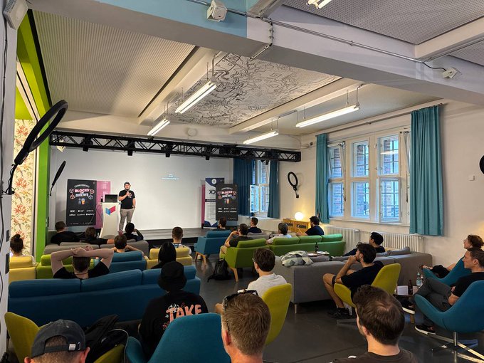 A presenter addresses a seated audience in a cozy lounge setup with sofas and armchairs, with banners reading "BLOCKCHAIN" visible beside the stage. The setting reflects the casual networking atmosphere described, bringing together speakers from XDC Network, PlatformD, SQD, Nillion, and Sphere Labs for Telekom & Friends Vol.3 in Berlin.