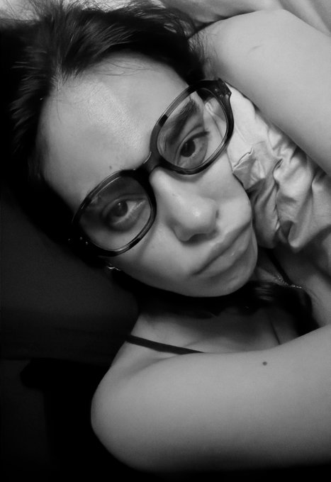 A black and white close-up portrait of a young woman with dark hair and large glasses lying down, gazing at the camera.