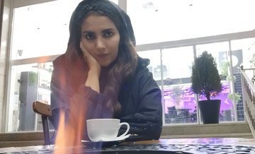 A woman with long dark hair sits at a table with a white coffee cup, resting her chin on her hand in what appears to be a cafe.