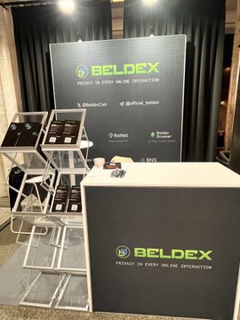 The image shows a booth for a company called Beldex at a trade show or event. The booth features a display of black and white posters advertising the company's services, including "Beldex" written in green letters. There are also several books on the table next to the posters. A small desk is present with a computer monitor and keyboard, suggesting that the company may be showcasing their online privacy solutions or providing information about them. The booth appears to be located near a window, allowing natural light to illuminate the display.