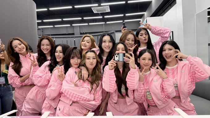 A large group of women wearing matching pink tracksuits pose cheerfully together, many flashing peace signs while one takes a mirror selfie with a smartphone, fitting the caption's excitement about waiting for an airdrop.
