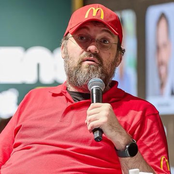 The image features a man wearing a red shirt with a microphone, suggesting that he might be speaking at an event or participating in a live stream. The background includes a green wall and a person's face, which could potentially be related to the post about Charles Hoskinson warning about DeFi apps shutting down.

In the context of the image, it seems like there is some sort of discussion or presentation taking place, possibly involving Charles Hoskinson's concerns about the future of DeFi applications. The man in the red shirt and microphone could be addressing these concerns directly to an audience or participating in a panel discussion.