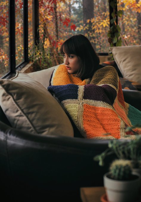 A young woman with dark hair sits wrapped in a colorful, patchwork crocheted blanket on a couch, gazing thoughtfully out a large window at an autumn forest.
