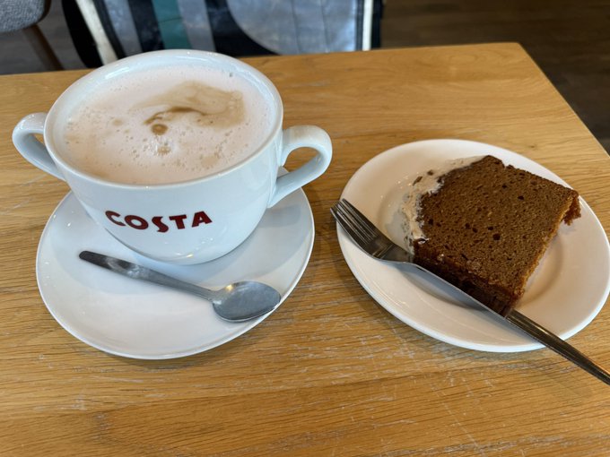 A Costa coffee cup filled with a frothy beverage sits on a saucer with a spoon, next to a plate holding a slice of cake on a wooden table.