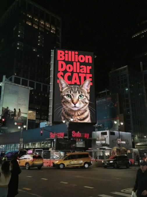 A nighttime street scene in Times Square featuring a large illuminated billboard that reads "Billion Dollar SCATE" above an image of a cat, with yellow taxis and a Subway restaurant visible below.