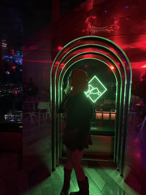 A woman stands in front of a glowing green neon arched mirror in a dimly lit, red-hued bar, with her reflection showing a diamond-shaped neon sign behind her.
