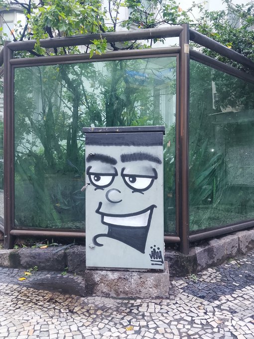 A grey utility box on a cobblestone sidewalk features a large, expressive face drawn in black graffiti, complete with a mustache and a wide grin.