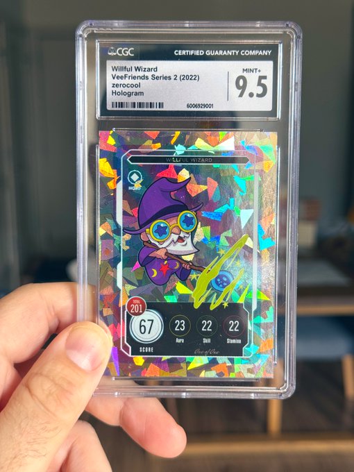 In the image, a person's hand is holding up an item that resembles a card or token from a trading card game. The player has just acquired this card, which they are showing off or possibly displaying for others. The card appears to be part of the @veefriends TCG collection and features a character with unique attributes like glasses and an odd hat. It's likely being discussed in relation to the post where it was shared or seen on social media platforms.