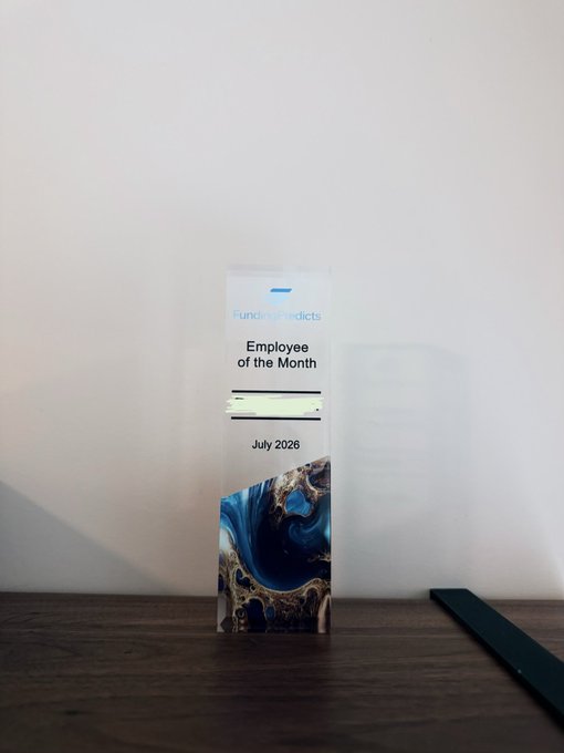 A clear, rectangular acrylic award or bookmark from 'Funding Predicts' sits on a wooden surface, engraved with 'Employee of the Month' and 'July 2026' above a blue and gold resin design.