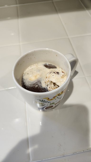 A white ceramic mug with a floral pattern sits on a white tiled counter, containing what appears to be ice cream or a dessert dissolving in a dark liquid, with a shadow cast across the tiles.
