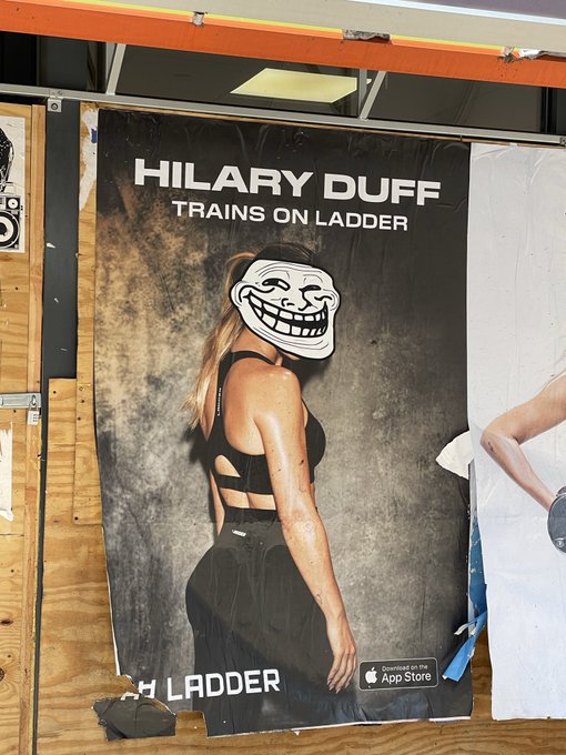 A fitness poster showing a woman in black workout attire with a troll face meme covering her head, titled 'HILARY DUFF TRAINS ON LADDER' promoting a workout app available on the App Store.
