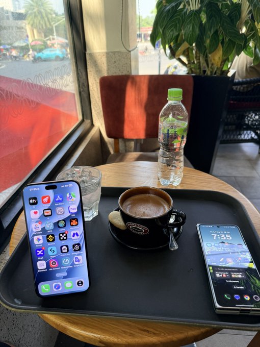 A first-person view at a café table showing a tray with two smartphones (an iPhone and a Samsung Galaxy), a black coffee cup, a glass of water, and a bottle of water, with a window view in the background.