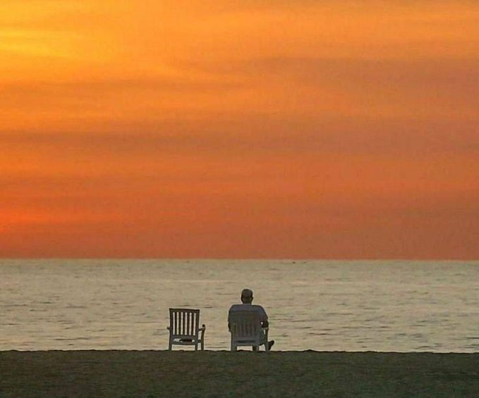 A serene photograph of a person sitting alone in a chair on a beach, watching an orange sunset over the ocean, with an empty chair beside them.