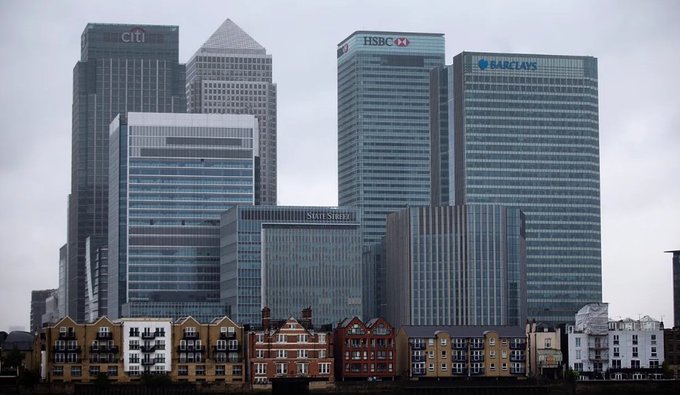 A view of the Canary Wharf skyline in London features skyscrapers labeled with logos for Citi, HSBC, Barclays, and State Street, with colorful residential buildings in the foreground.