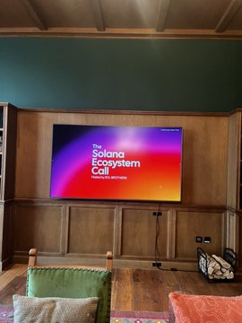 A living room with wood-paneled walls featuring a TV displaying 'The Solana Ecosystem Fall 2024' screen, with green and orange pillows on a chair below.
