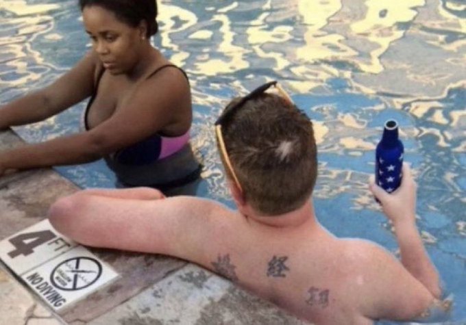 The image shows a man with tattoos sitting in a pool of water next to another person who appears to be holding a beer bottle. The scene takes place near a sign that says "4F". The man has tattoos on his back, which are visible from this angle. The woman beside him is wearing a bathing suit and sunglasses. They seem to be enjoying their time together in the pool area.