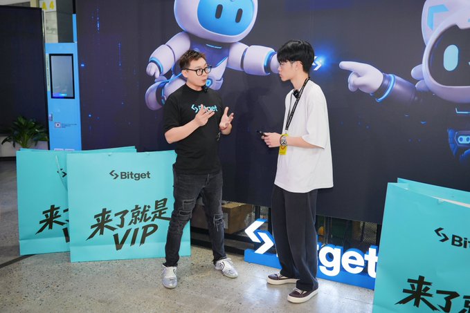 Two men converse at a Bitget promotional event with a robot-themed backdrop; one wears a black Bitget t-shirt, and turquoise shopping bags with the text '来了就是VIP' are visible.