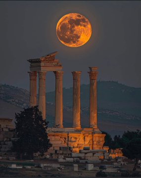 A scenic photograph of ancient stone columns at a Roman ruin illuminated at night with a large, glowing orange full moon hanging in the dark sky above.