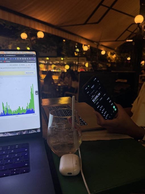 A nighttime scene at an outdoor café where someone holds a smartphone displaying a stock chart with the number '52,696.34,' next to a laptop showing a similar chart and a glass of ice water.
