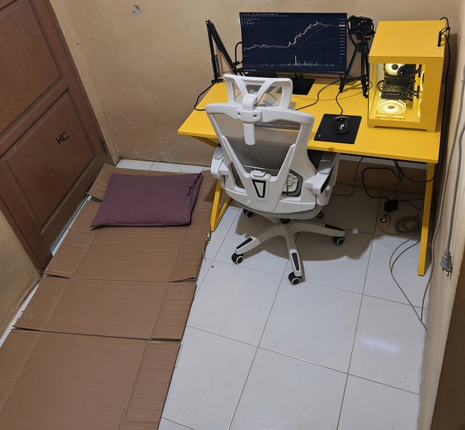A minimalist home office setup featuring a bright yellow desk, white ergonomic chair, and trading monitor displaying a rising financial chart, set on a bare tile floor with only a flat cardboard mat and pillow for floor seating. The sparse, no-frills arrangement embodies the Spartan ideal of rejecting luxury and comfort, reinforced by the #Locked hashtag suggesting the trader willingly endures a harsh environment to build discipline and strength.