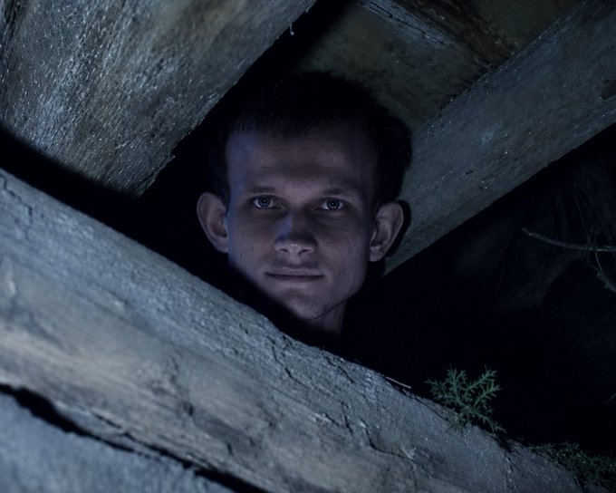 A young man with short hair peers out from between weathered wooden boards in a dark, shadowy setting illuminated by cool blue light.