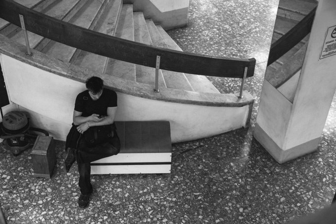 The image shows a man sitting alone on a bench, looking down at his hands. The bench appears to be located near stairs or a staircase, as there are steps visible in the scene. The photo has been edited with black and white tones, giving it an artistic touch. The caption provided suggests that this image is related to a post about taking breaks during the day.