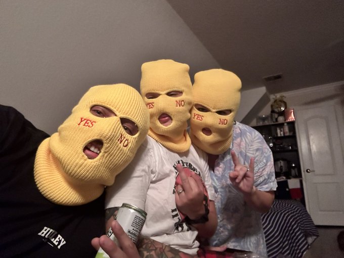 Three people posing indoors while wearing yellow balaclava ski masks with "YES" and "NO" printed in red over the eye and mouth holes.