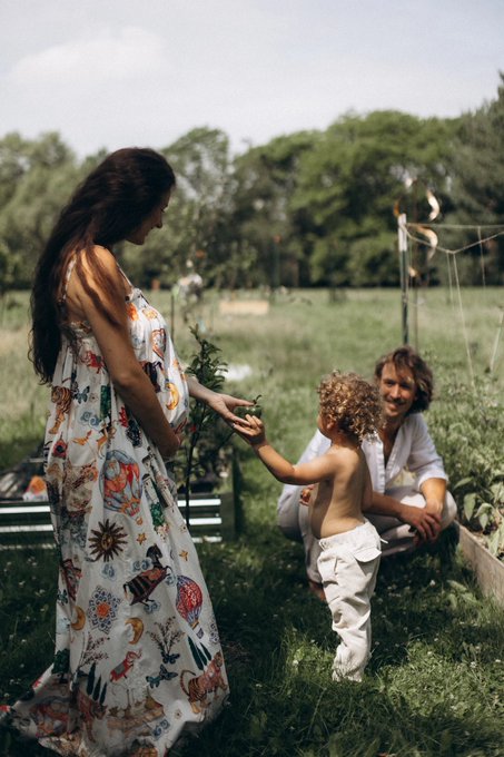 A woman in a floral maxi dress hands a plant to a curly-haired toddler, while a smiling man kneels beside him in a sunlit garden. The scene captures the simple joy of a family gardening together, evoking the peaceful escape implied by "Mentally I'm here."