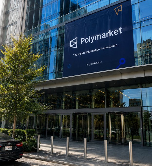 Exterior photo of a modern glass office building displaying a large banner that reads 'Polymarket - The world's information marketplace' with the website polymarket.com.