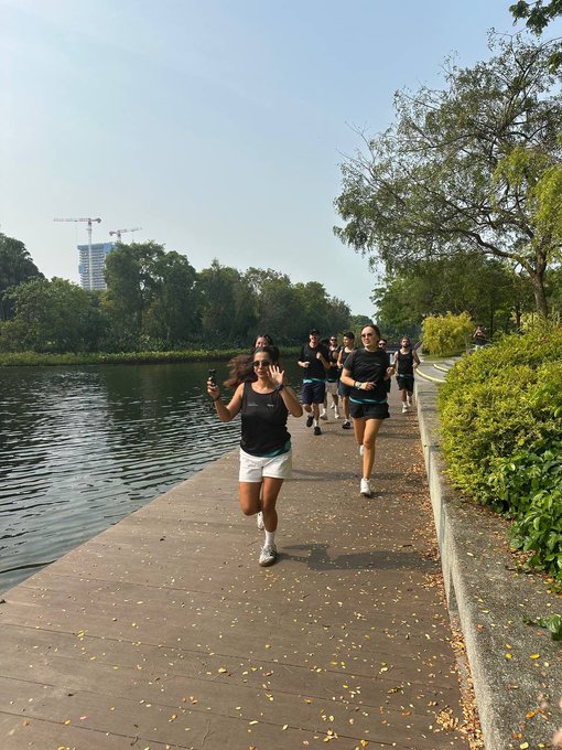 A morning group of runners, including a woman waving at the camera, jog along a lakeside path at Marina Bay with construction cranes visible in the background, illustrating the Fhenix team's sunrise run where their fitness data stayed private thanks to the Hyme app as they celebrated Fhenix mainnet's October launch.