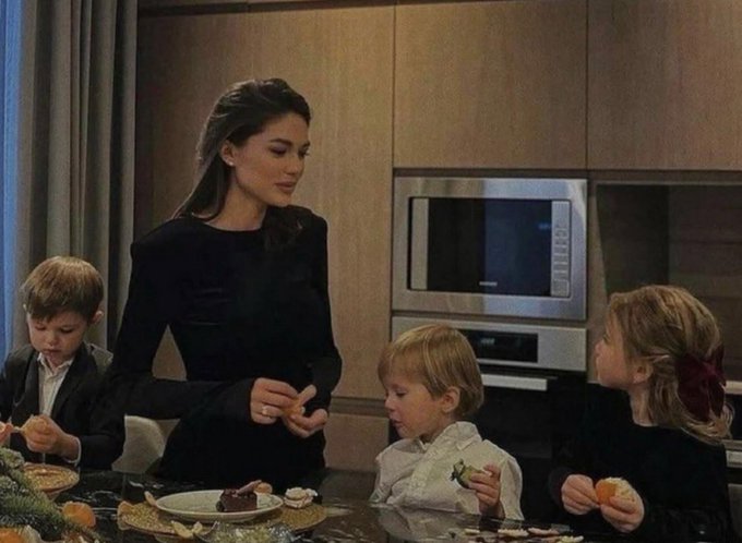 A woman in a black dress serving food to three young children at a kitchen counter in a modern home.