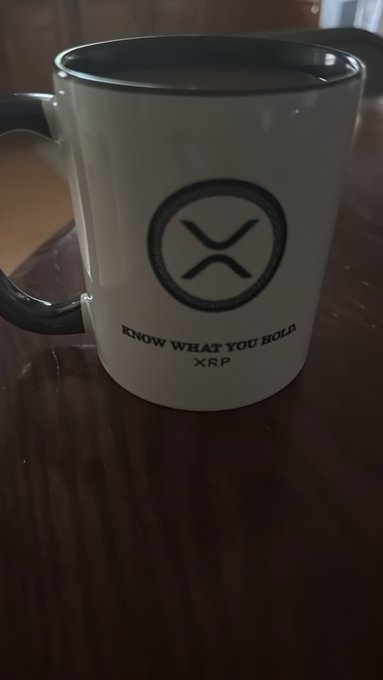 A white ceramic mug with black rim and handle featuring the XRP (Ripple) logo and the text 'KNOW WHAT YOU HOLD XRP' sitting on a dark wooden surface.