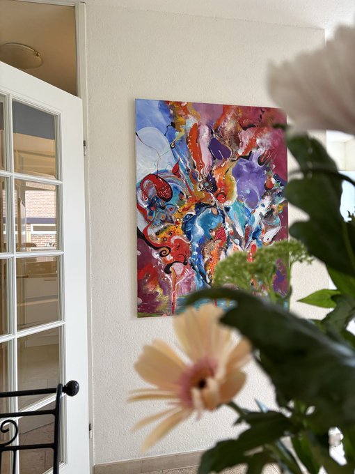 A vibrant, colorful abstract painting hangs on a white wall beside a glass-paned door, with a pale pink flower blurred in the foreground.