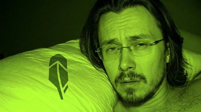 A close-up of a bearded man with glasses lying on a pillow, with the entire image tinted green and a green logo resembling a stylized arrow or shovel next to him.