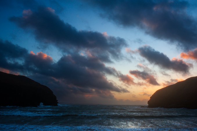 A scenic seascape at sunset or sunrise with dramatic pink-tinged clouds over dark silhouetted cliffs and waves rolling into the ocean.