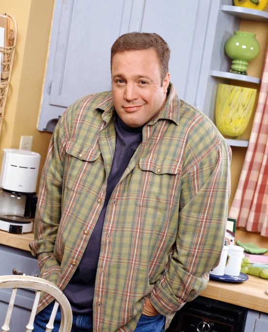 A man in a green and brown plaid shirt over a dark t-shirt stands smiling in a kitchen set with yellow walls, blue cabinets, and a green vase.