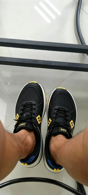 First-person view looking down at a person wearing black and yellow sneakers with distinctive diamond-shaped logos on the tongue, standing on a glossy floor between black metal rails.