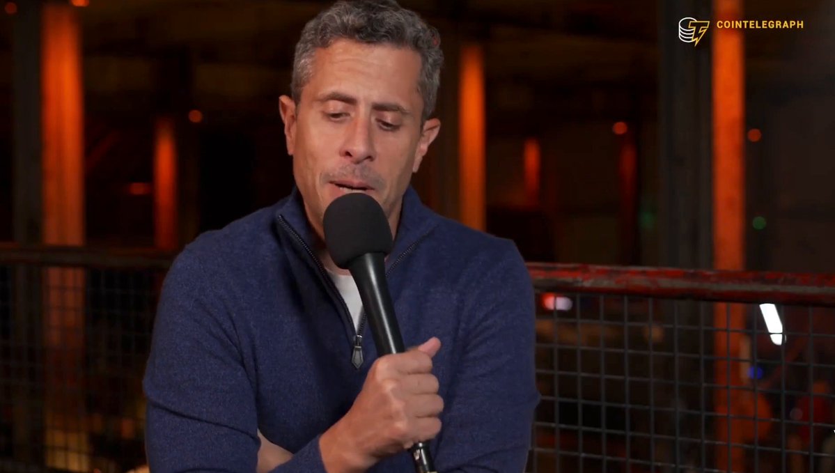 The image shows a man wearing a blue sweater holding a microphone, possibly speaking into it or recording something. He appears to be in front of a fence with a building behind him. There are also other people visible in the background, but they do not seem to be the main focus of the scene.
