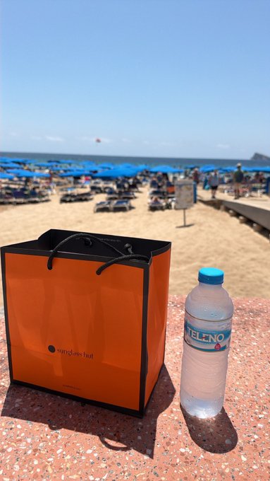 An orange "sunglass hut" shopping bag and a plastic bottle of "Vittel" water rest on a stone surface, with a crowded beach, blue umbrellas, and the ocean visible in the background.