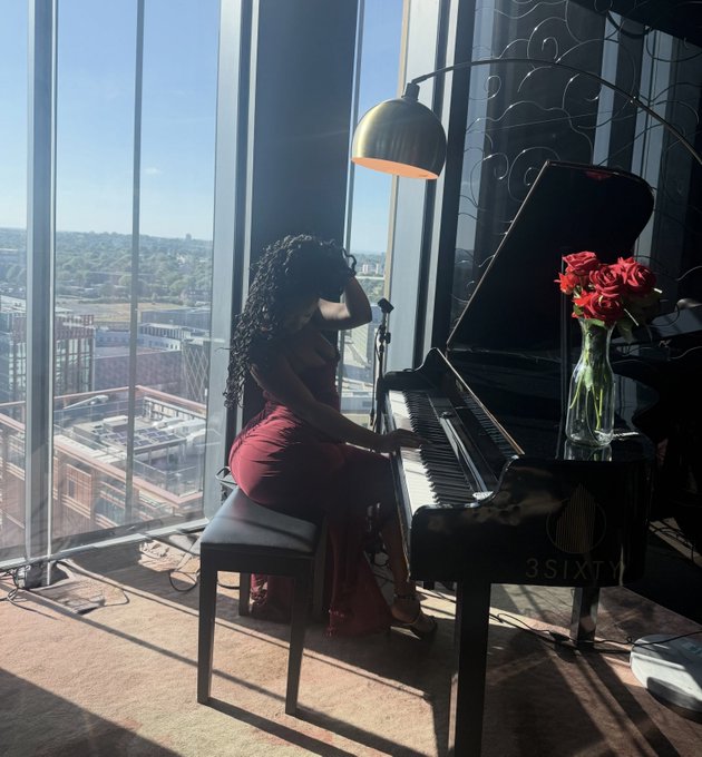 A woman with long curly hair in an elegant red dress sits at a grand piano by large floor-to-ceiling windows overlooking a cityscape, with a vase of red roses beside her and a '3SIXTY' watermark visible.