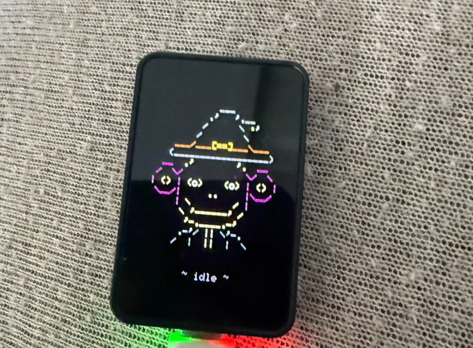 A small black handheld electronic device displaying an animated pixel character wearing a witch hat, with the text '~ idle ~' on the screen, sitting on a mesh surface.