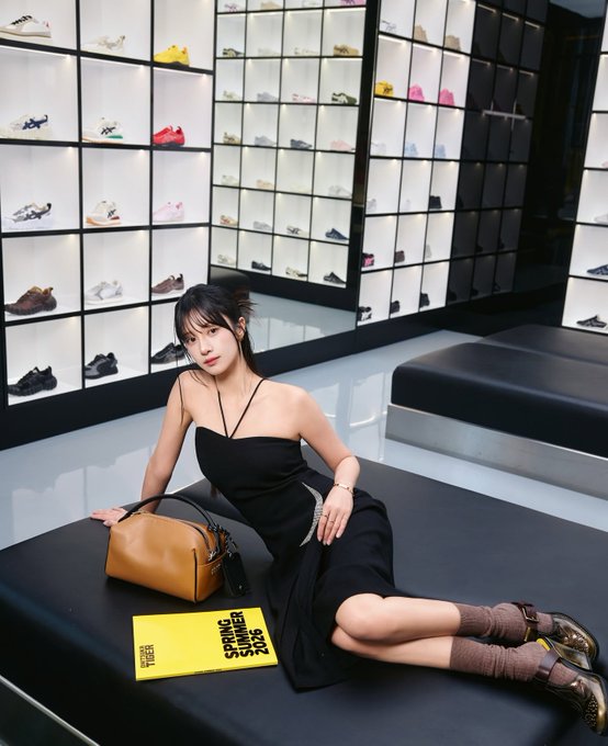 A young woman in a black dress reclines on a dark bench in a room filled with wall-to-wall shelving displaying various sneakers. A yellow magazine titled 'HIGH SNOBIETY SPRING 2023' and a tan handbag sit beside her.