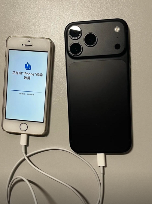 An older iPhone connected to a newer black iPhone via a charging cable, with the screen displaying Chinese text "正在向iPhone传输数据" (Transferring data to iPhone) with a progress bar.