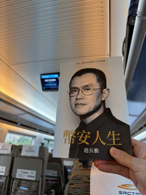 A hand holding a Chinese book titled '常安人生' (Cháng'ān Rénshēng) by author 赵长鹏 (CZ, Changpeng Zhao), photographed inside what appears to be a high-speed train.